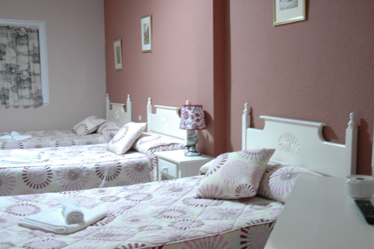 Habitación de hotel con dos camas individuales, cada una con cabecera blanca y almohadas a juego. Se aprecia una mesita de noche con lámpara y un cajón pequeño. Las paredes están pintadas de un rosa suave, y hay cuadros enmarcados.