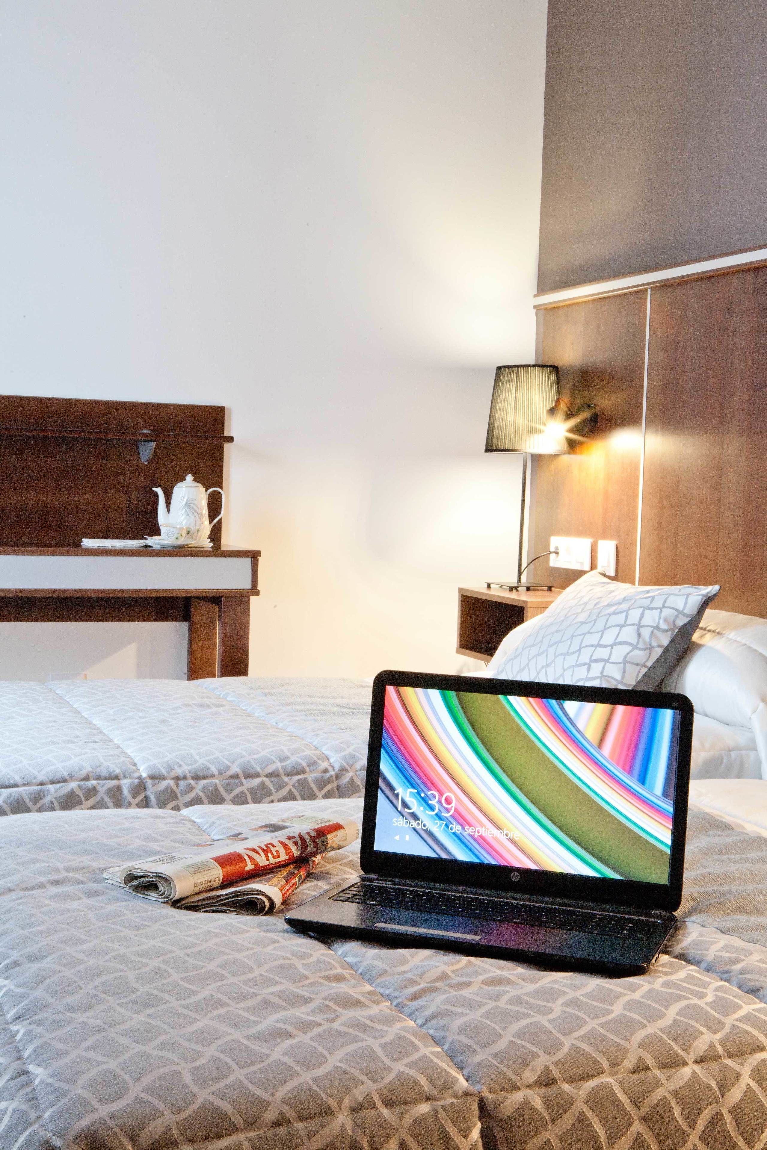 A hotel room with a bed, a nightstand, a lamp, a laptop, and a newspaper.