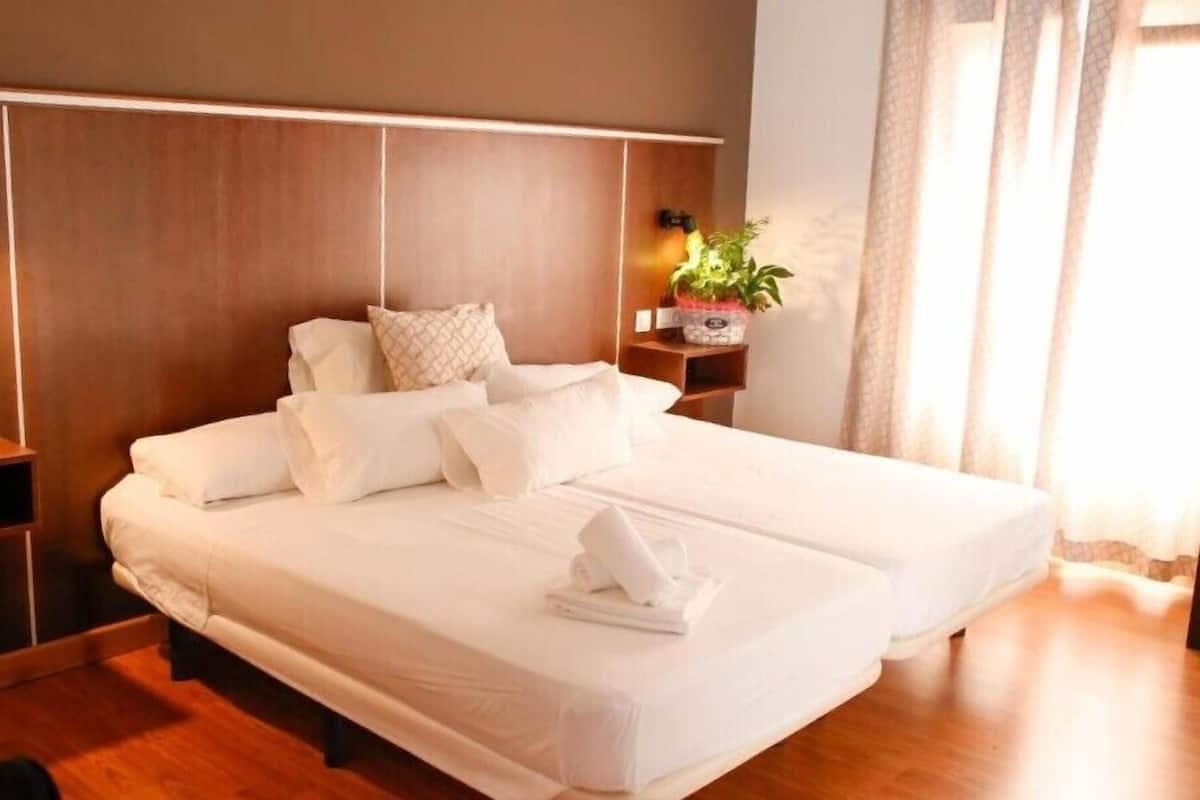 Una cama doble con sábanas blancas y una toalla doblada sobre la cama. Hay una mesita de noche con una lámpara y una planta en maceta al lado de la cama.