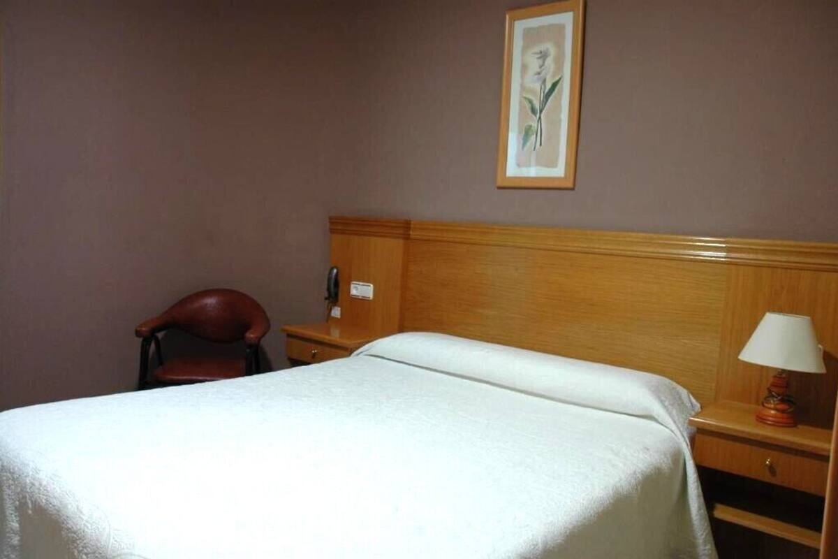 Una habitación de hotel con una cama individual, cabecera de madera, una silla, una mesita de noche con lámpara y un cuadro en la pared.