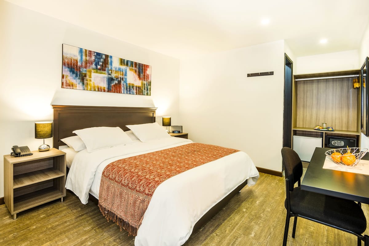 Una habitación de hotel con una cama grande, un escritorio con silla, una mesita con frutas y un cuadro en la pared.