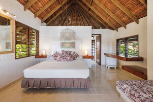 A spacious bedroom with a large bed, wooden ceiling beams, and a view of greenery through the windows.