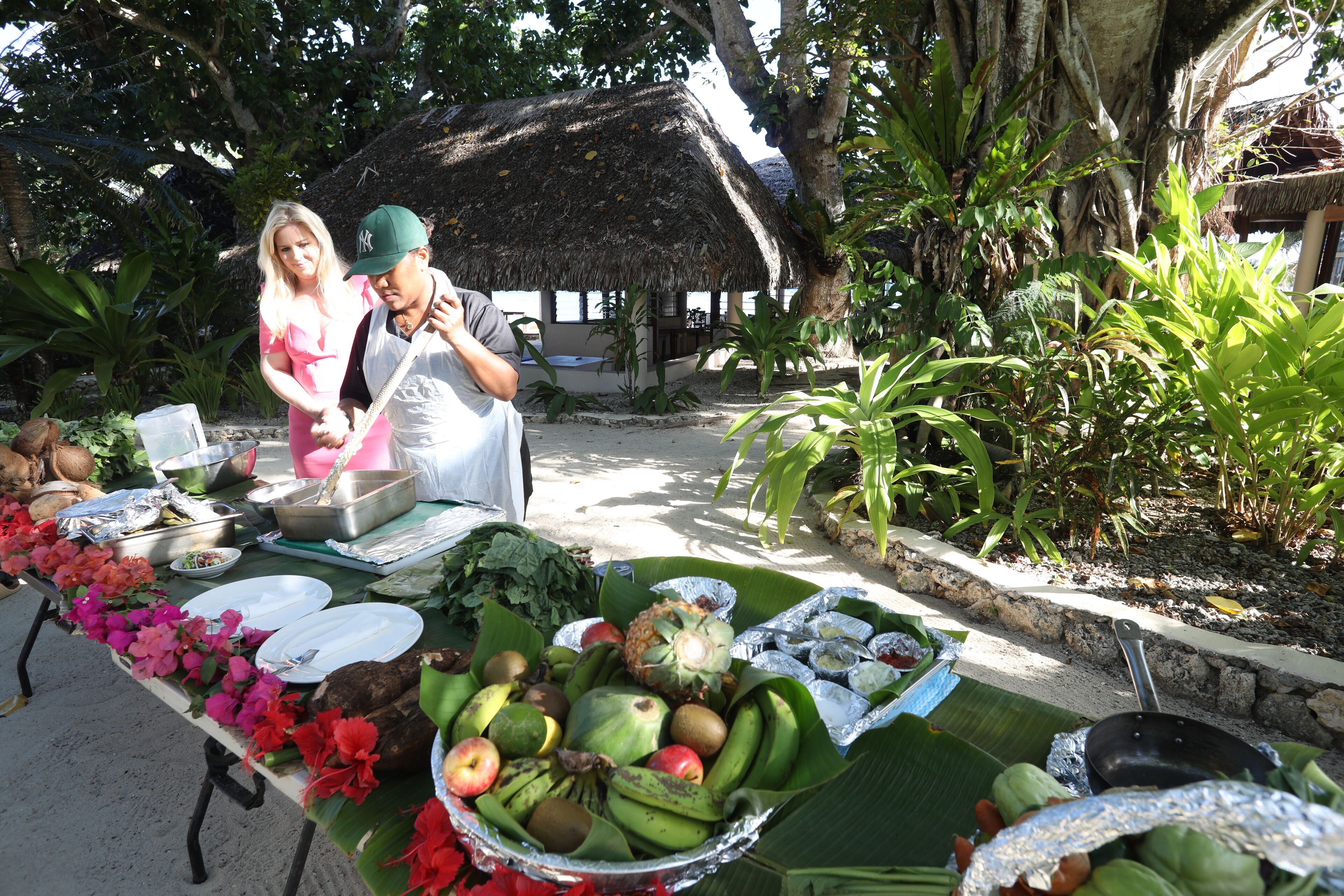 Un área de comedor al aire libre con ambiente tropical, que cuenta con una estructura de techo de paja, diversos frutos y verduras sobre una mesa, y una persona de pie cerca.
