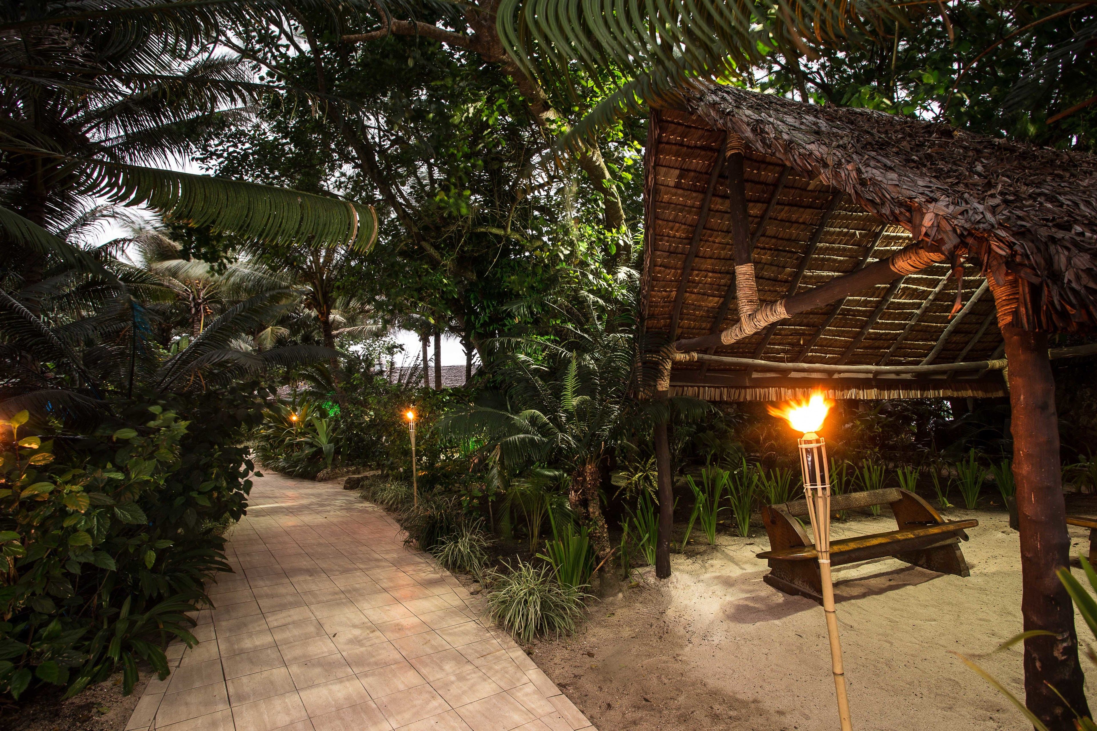 A pathway leading to a thatched-roof structure with a bench and a torch.