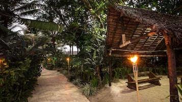 A pathway leading to a thatched-roof structure with a bench and a torch.