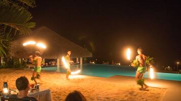 A nighttime beach scene with a pool, palm trees, and a thatched-roof structure.