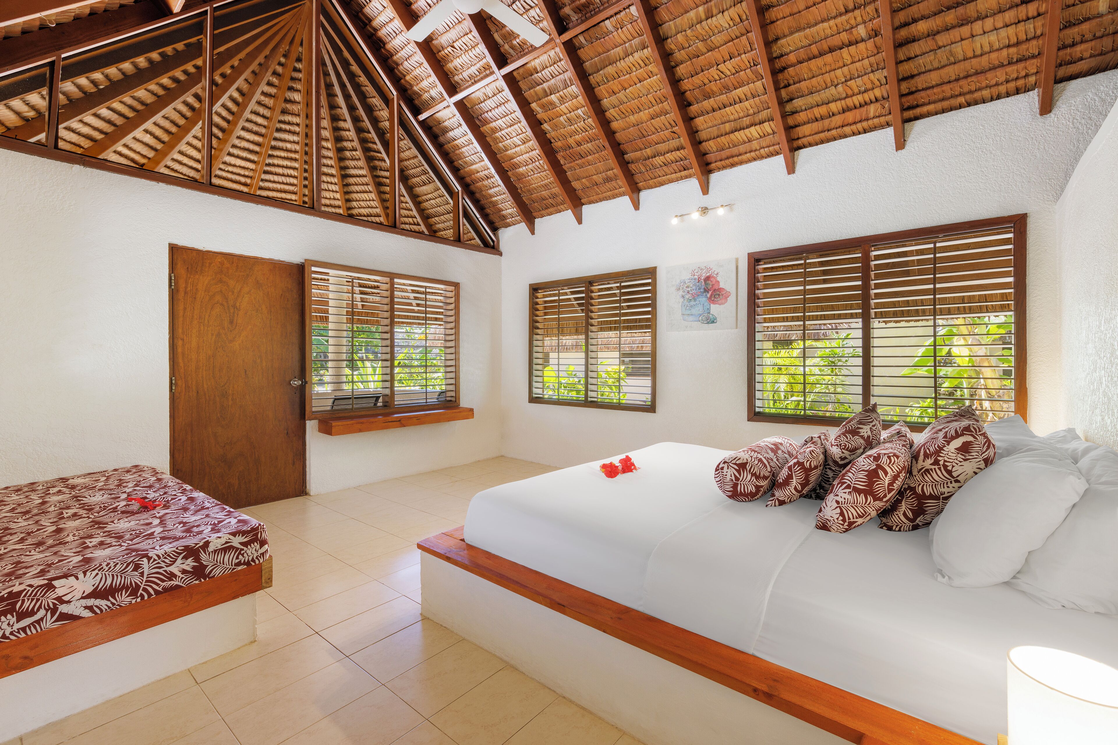 Un dormitorio con techo de madera, una cama con almohadones estampados, una ventana con persianas y una puerta.