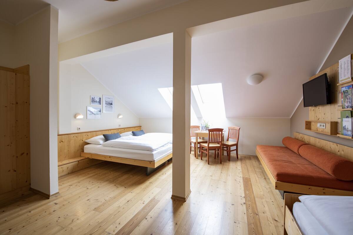Una habitación con cama elevada, un sofá, una mesa de comedor con sillas y un televisor montado en la pared.