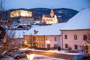 Un pueblo cubierto de nieve con edificios históricos, incluyendo una iglesia con una alta aguja, y un castillo al fondo.