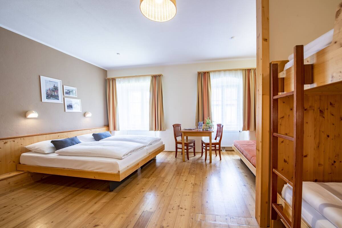 Una habitación con piso de madera, una cama con ropa blanca, una mesa con sillas y una cama nido.