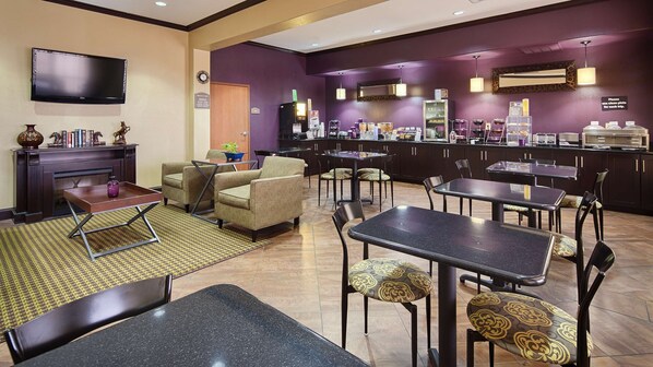A hotel lobby with a seating area, a fireplace, a television, a coffee station, and a counter with various food items.