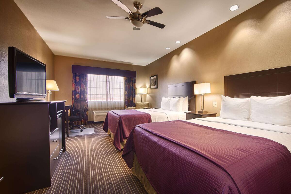 A hotel room with two beds, a desk with a television, a ceiling fan, and a window with curtains.