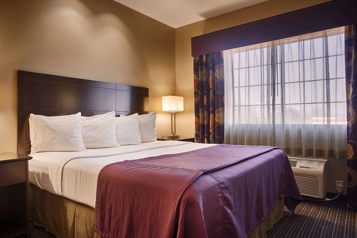 A hotel room with a large bed, two bedside tables, a window with curtains, and a wall-mounted air conditioning unit.