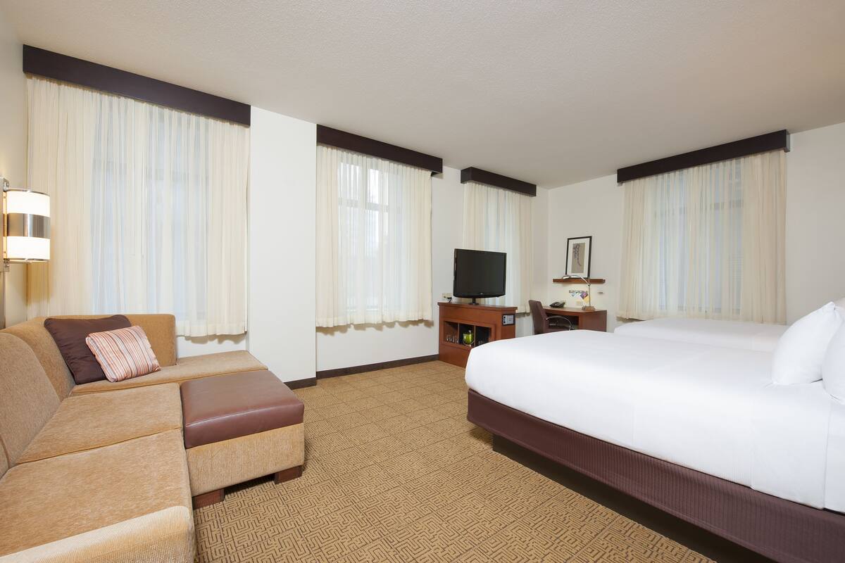 A hotel room with a large bed, a sofa, a TV on a wooden stand, and a small table with a lamp.