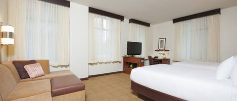 A hotel room with a large bed, a sofa, a TV on a wooden stand, and a small table with a lamp.
