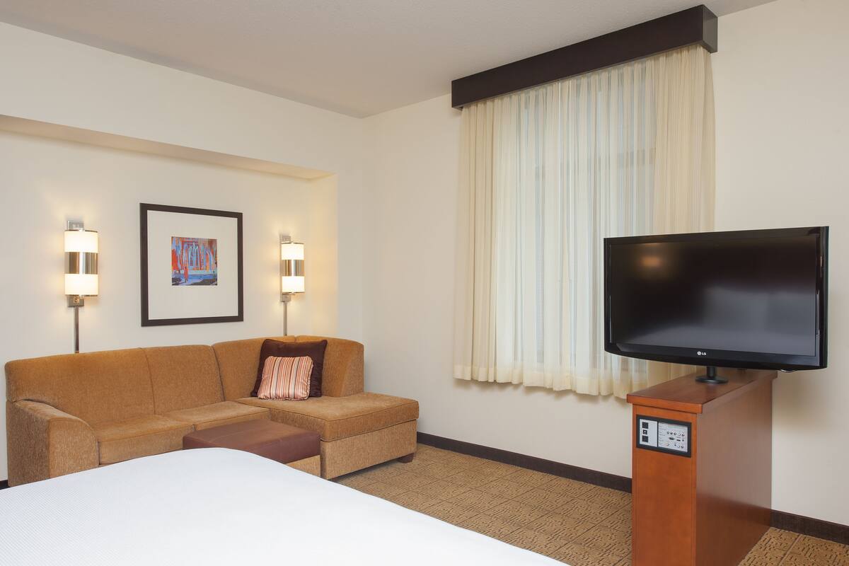 A hotel room with a bed, a sofa, a television on a wooden stand, and two wall-mounted lamps.