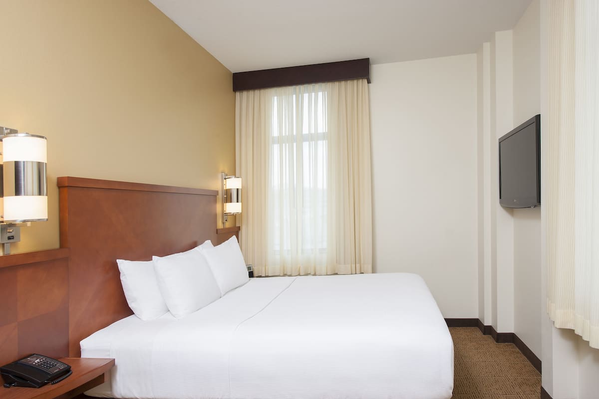 A hotel room with a large bed, a wooden headboard, a bedside table with a lamp, a flat-screen TV mounted on the wall, and a window with sheer curtains.