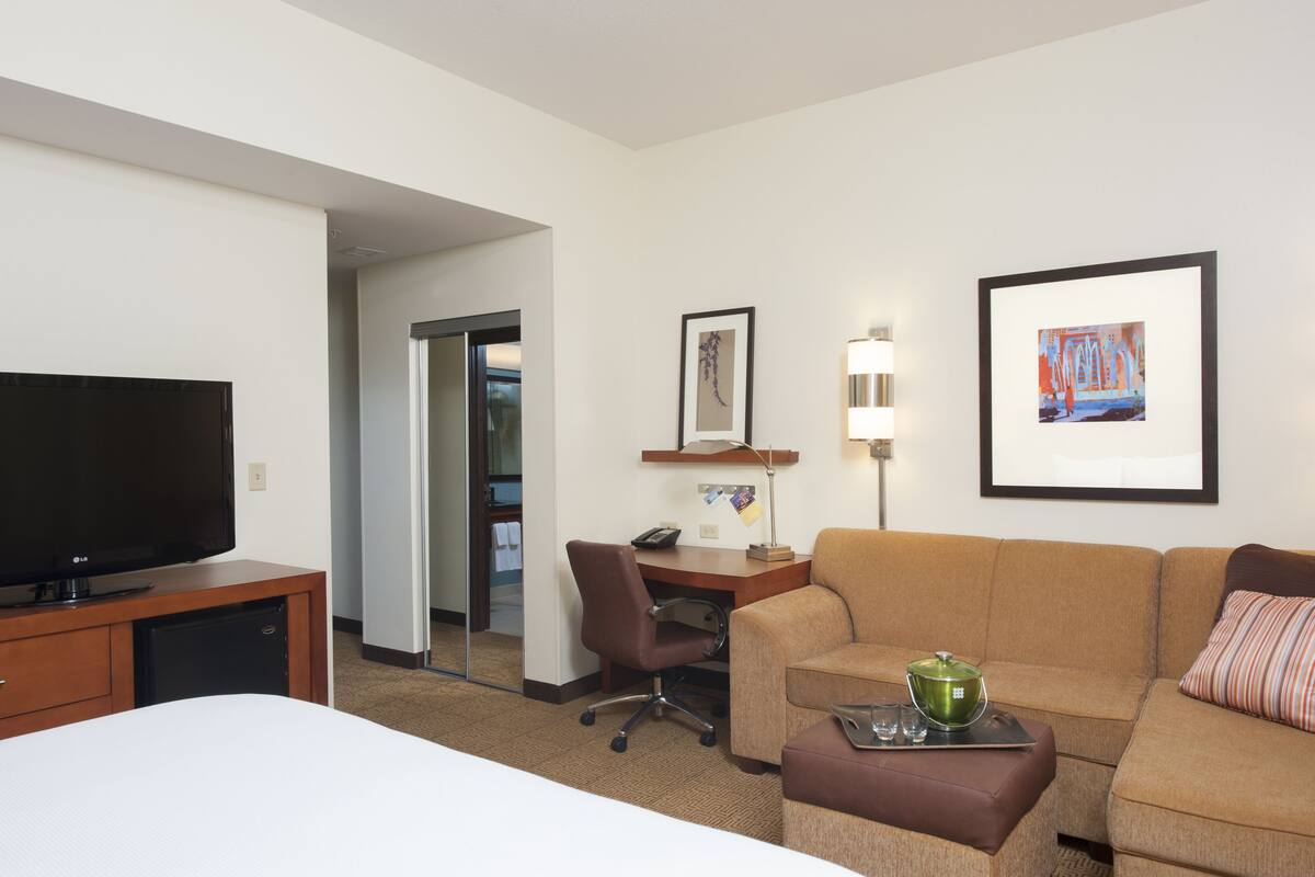 A hotel room with a bed, a TV, a desk with a chair, a couch, and a painting on the wall.