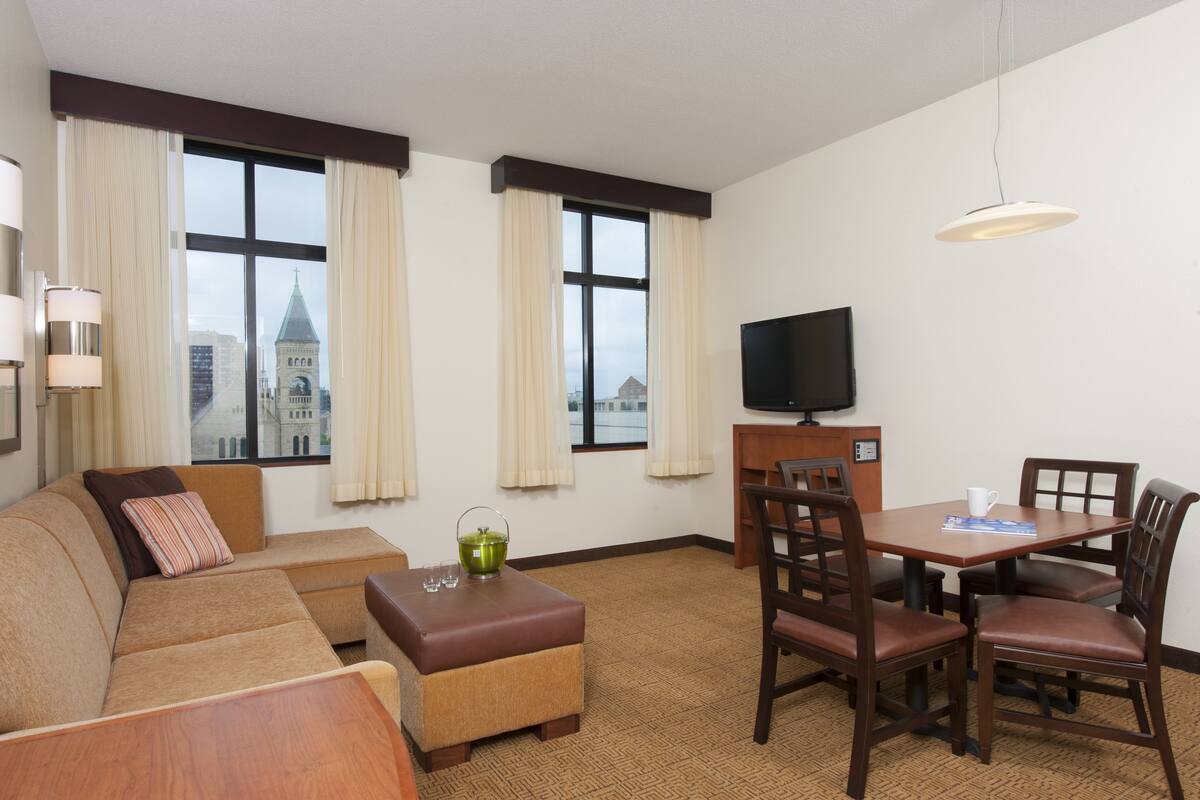 A hotel room with a sofa, ottoman, dining table with chairs, a television, and large windows with curtains.