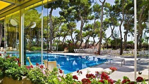 Un área de piscina con una piscina azul cristalina, rodeada de sillas y mesas blancas y abundante vegetación.