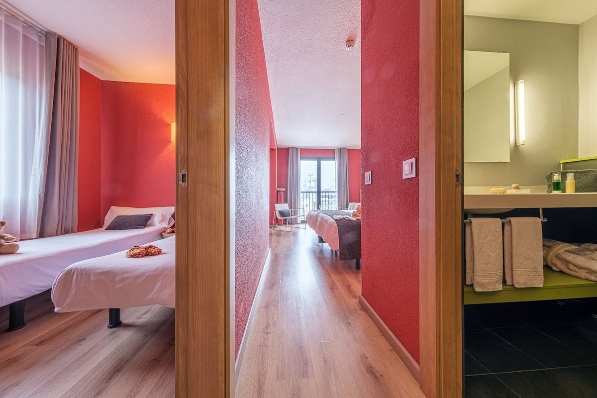 A hotel room with two beds, a bathroom with a mirror and towel rack, and a hallway leading to another room.