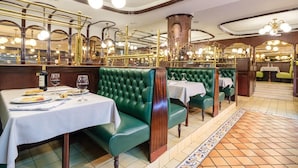 L’intérieur d’un restaurant présente des banquettes recouvertes d’un tissu vert, des nappes blanches et des luminaires en laiton décoratifs.