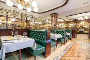 L’intérieur d’un restaurant présente des banquettes recouvertes d’un tissu vert, des nappes blanches et des luminaires en laiton décoratifs.