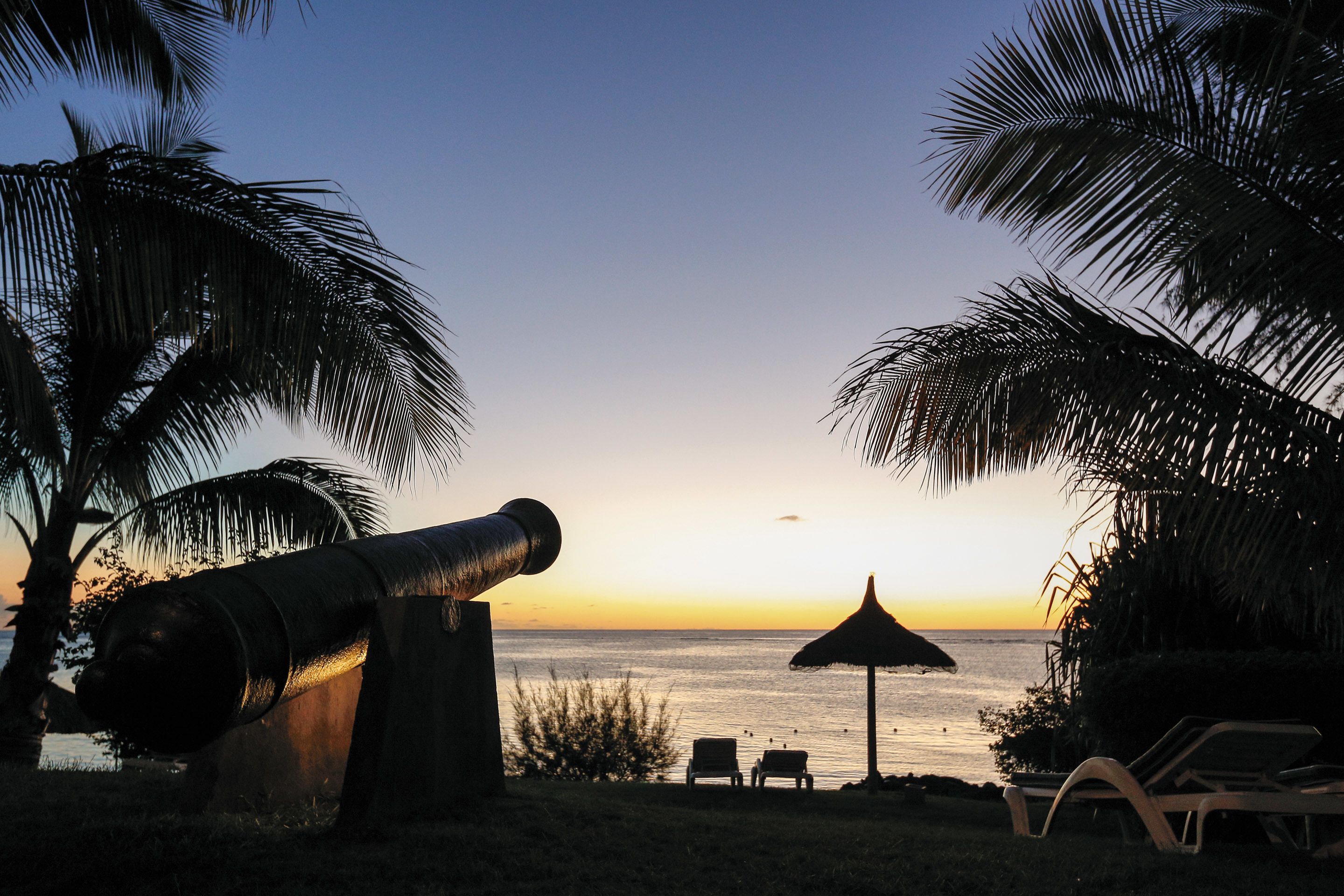 A beach scene with a cannon, lounge chairs, and a thatched umbrella.