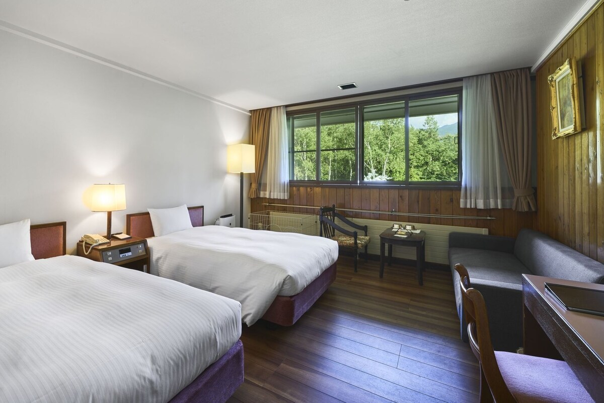 A hotel room with two beds, a desk, a chair, and a large window with curtains.