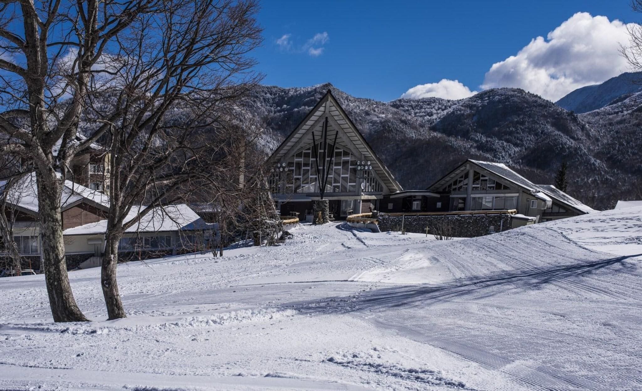 Een besneeuwd landschap met een chalet met een opvallend driehoekig dak en een skipiste op de achtergrond.