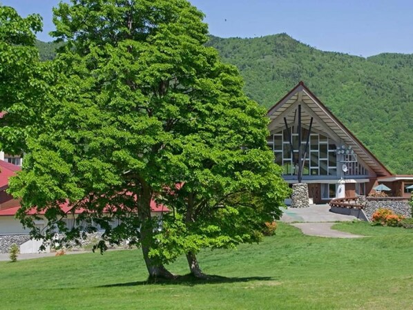 A lodge with a large tree in front, a mountain backdrop, and a modern building with a triangular roof. - Okushiga Kogen Hotel (Yamanouchi)