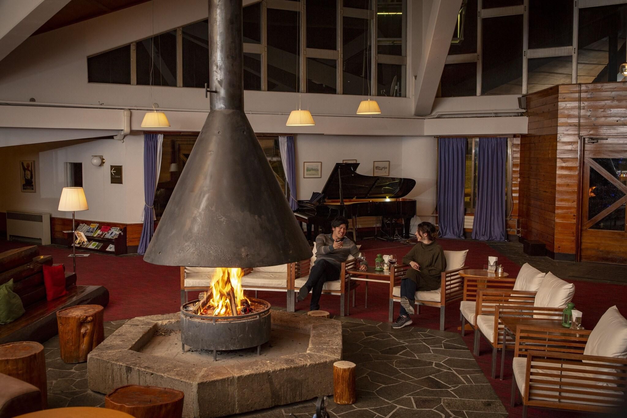 Een ruime lobby met een centrale houtkachel, comfortabele zitplaatsen, een vleugelpiano en grote ramen.
