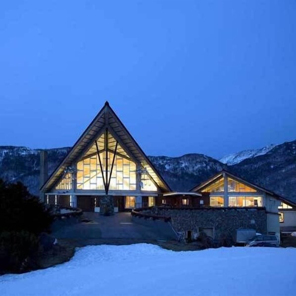 Um edifício grande, em formato triangular, com uma fachada de vidro central imponente, rodeado por neve e montanhas.