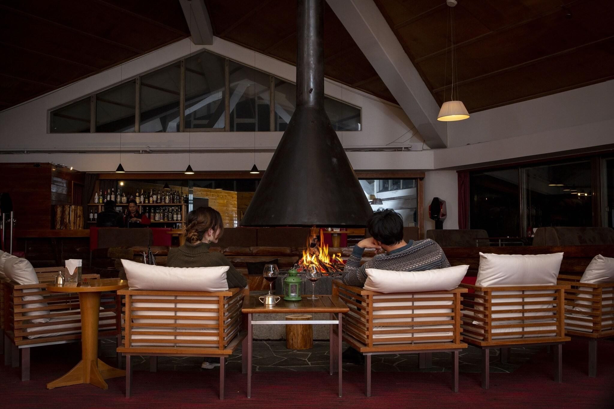 Een gezellig zithoekje met houten stoelen en een centrale tafel, met een grote, zwarte schoorsteen en een bar op de achtergrond.