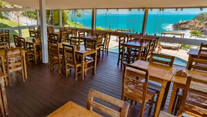 An outdoor dining area with wooden tables and chairs, overlooking a scenic ocean view. - Apa Pau Brasil (Búzios)