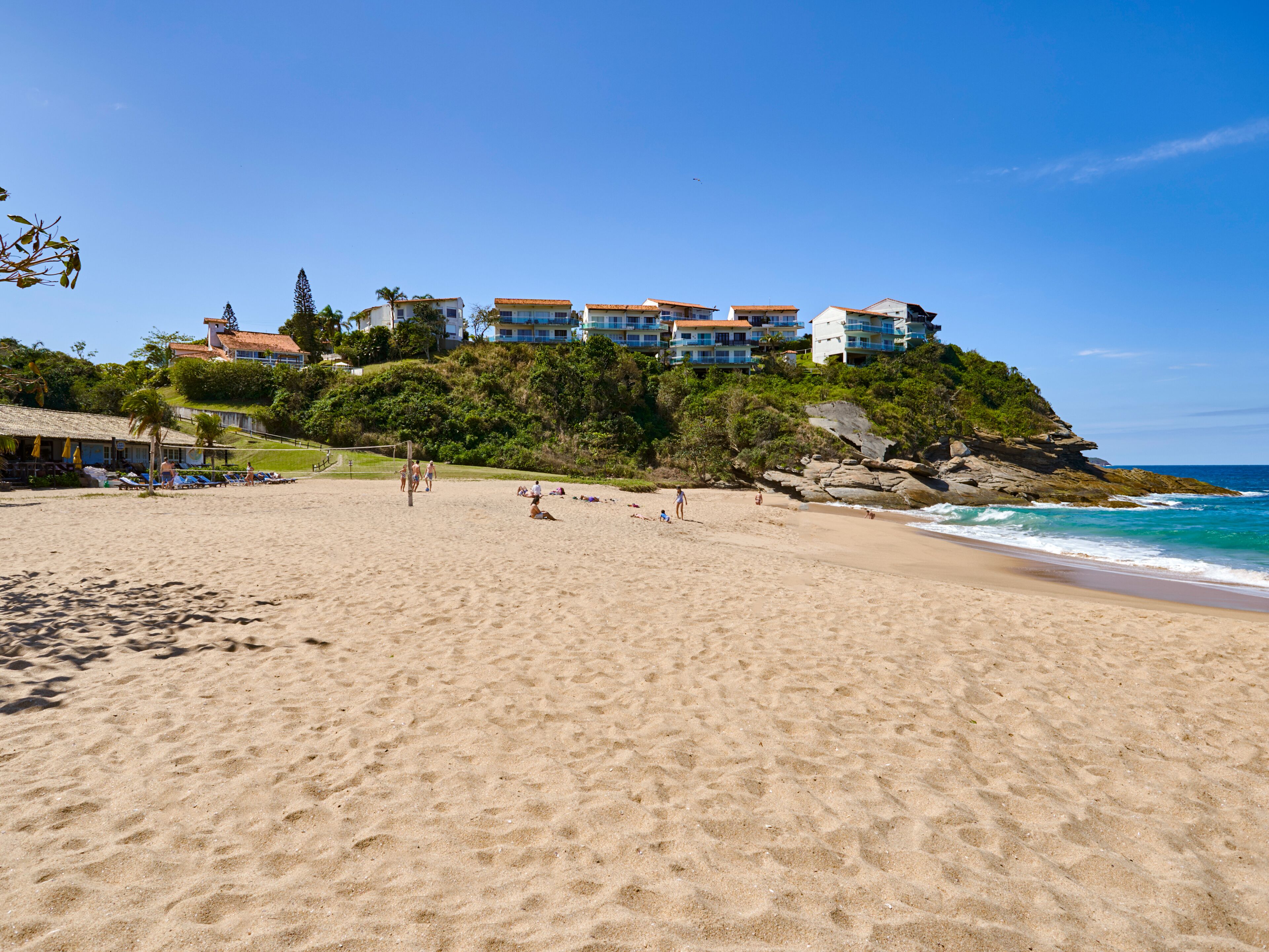 Uma praia com um edifício grande numa colina, céu claro e ondas do oceano.