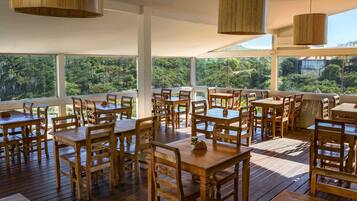 Un área de comedor al aire libre con mesas y sillas de madera, y ventanas amplias que ofrecen vistas de la vegetación circundante.