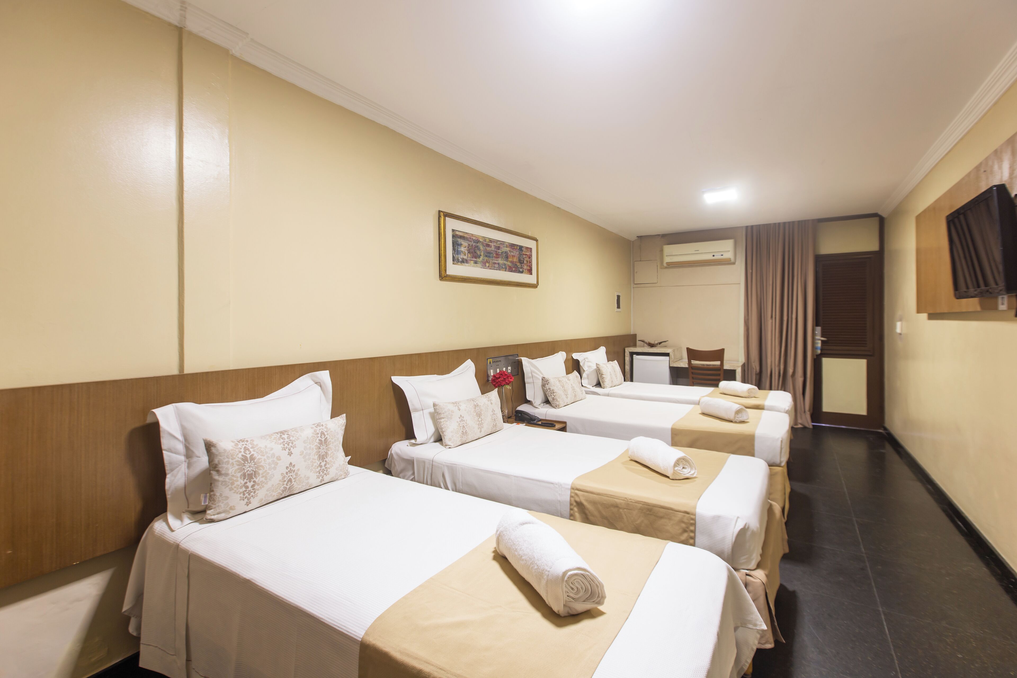 Habitación de hotel con cuatro camas individuales, un televisor de pantalla plana montado en la pared, un equipo de aire acondicionado y un cuadro enmarcado.