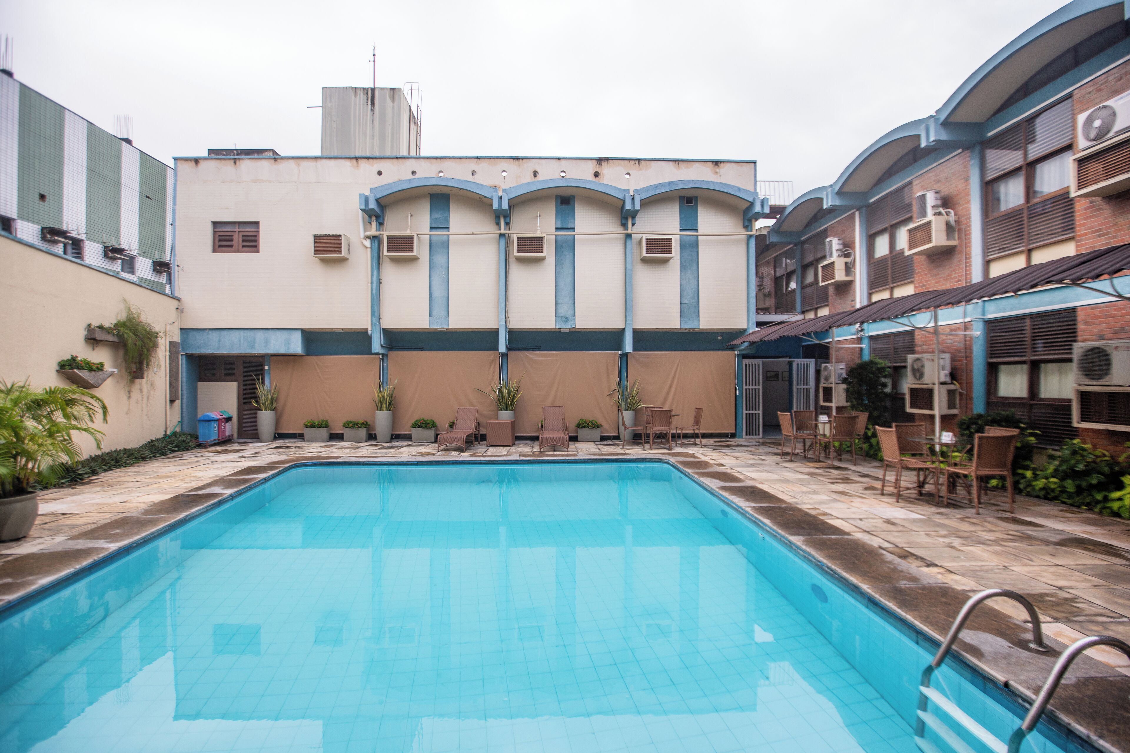 A swimming pool with lounge chairs and potted plants, surrounded by buildings with air conditioning units.