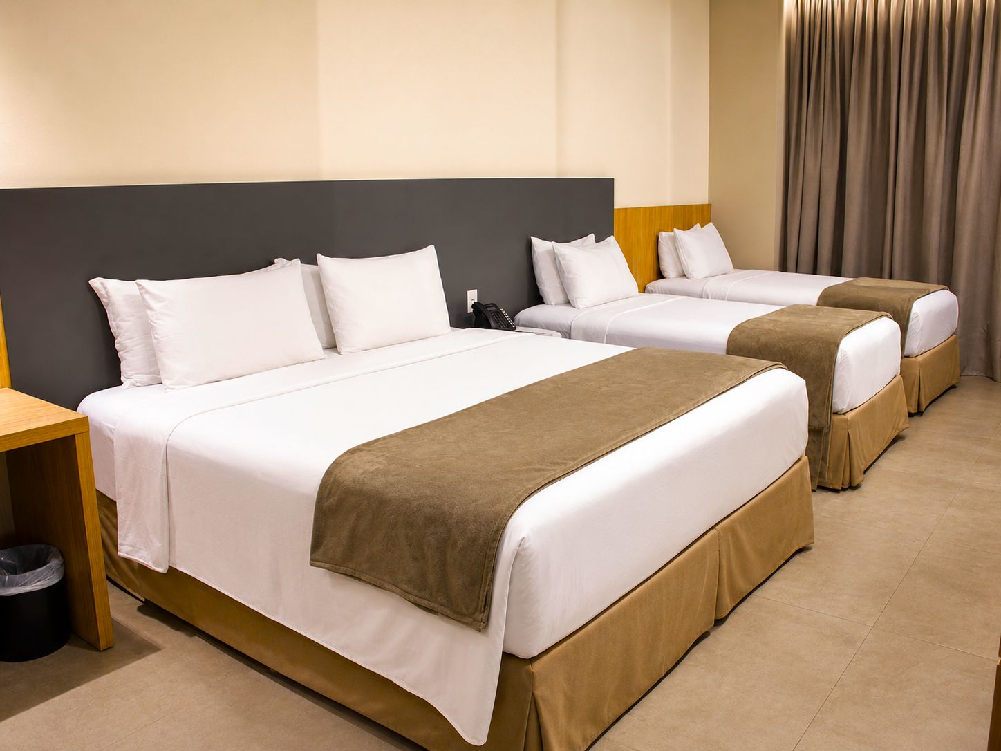 A hotel room with two beds, a wooden nightstand, a trash bin, and a curtain.
