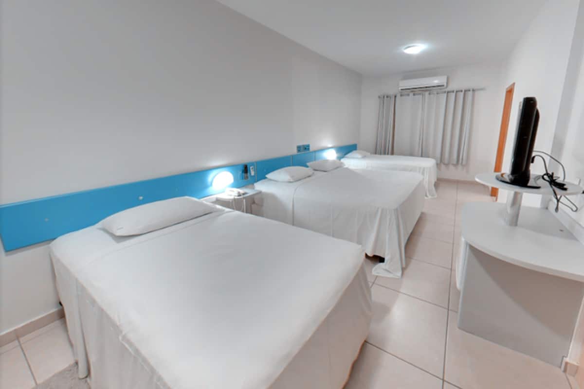 Une chambre d’hôtel avec trois lits simples, un petit bureau avec une chaise, une télévision et une salle de bain avec un miroir et un lavabo.