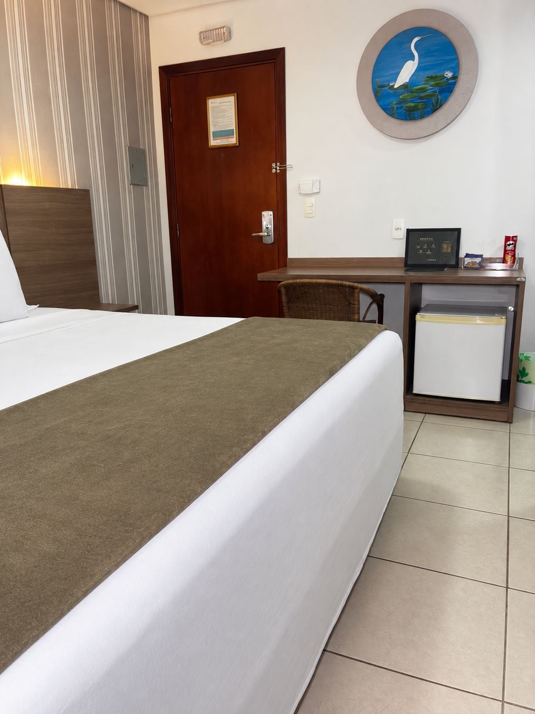 Habitación de hotel con cama, escritorio, silla, refrigerador pequeño y una obra de arte colgada en la pared.