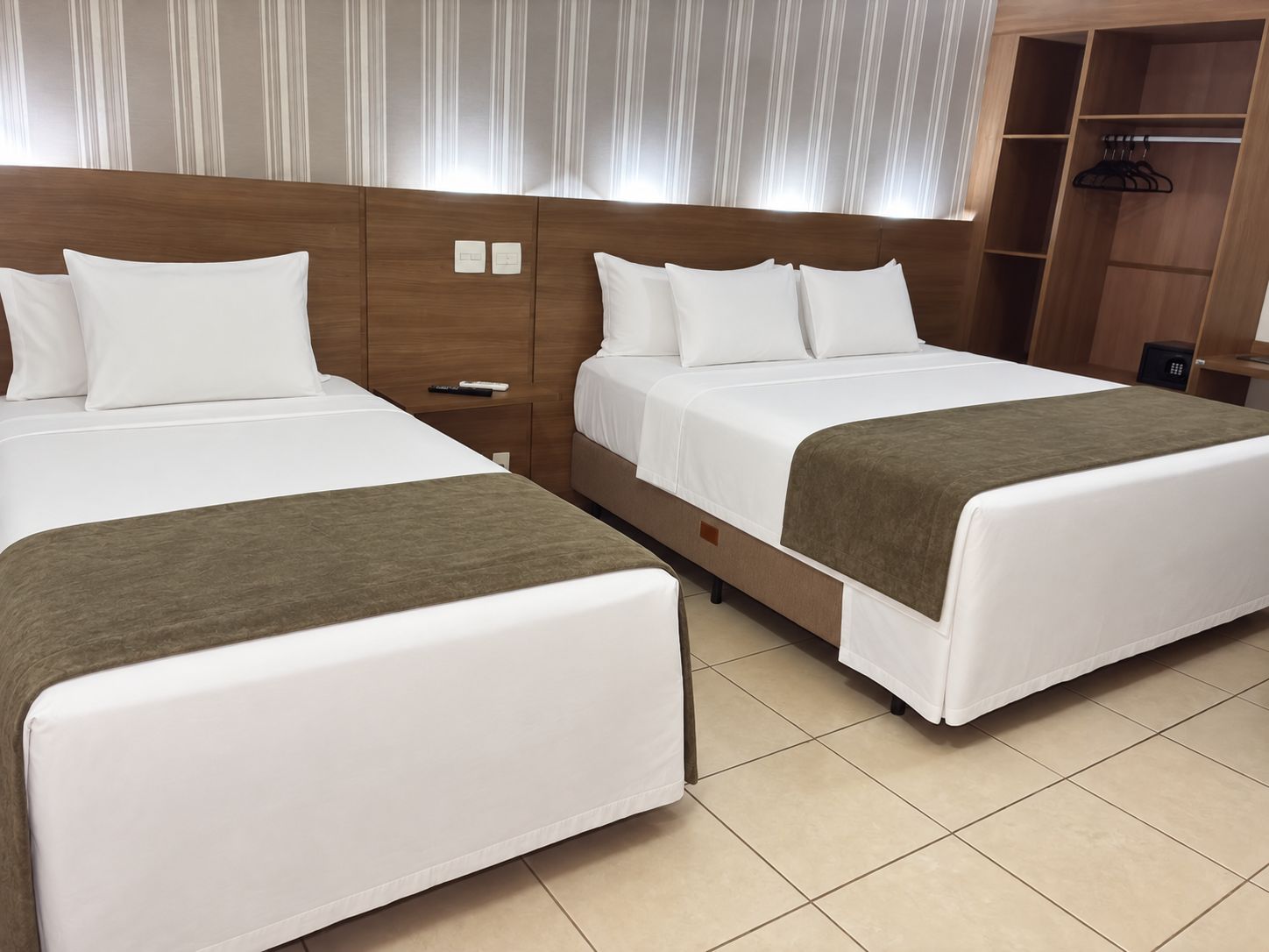 Quarto de hotel com duas camas, cabeceira de madeira, guarda-roupa e mesa de cabeceira.