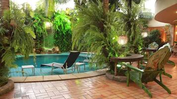 An indoor pool area with lounge chairs and tables, surrounded by lush greenery and palm trees.
