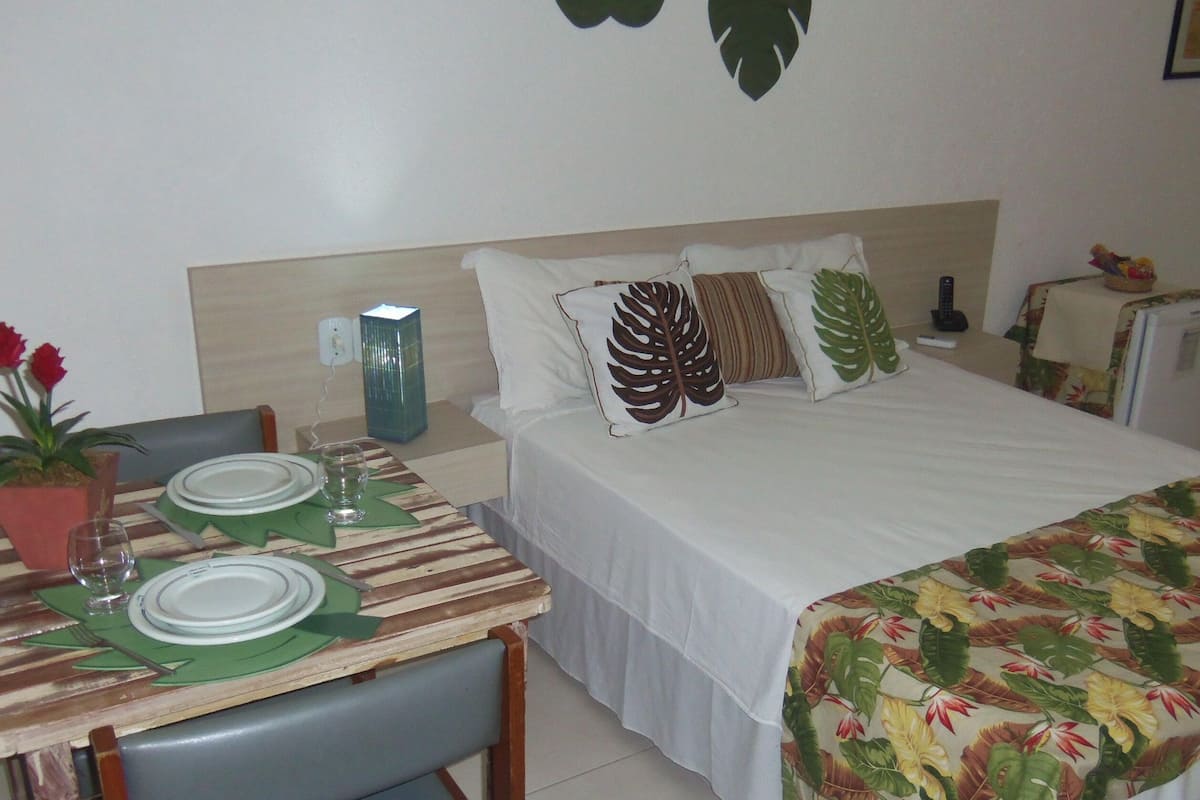 A hotel room with a bed, a bedside table, a small refrigerator, a table with a floral tablecloth, and a chair.