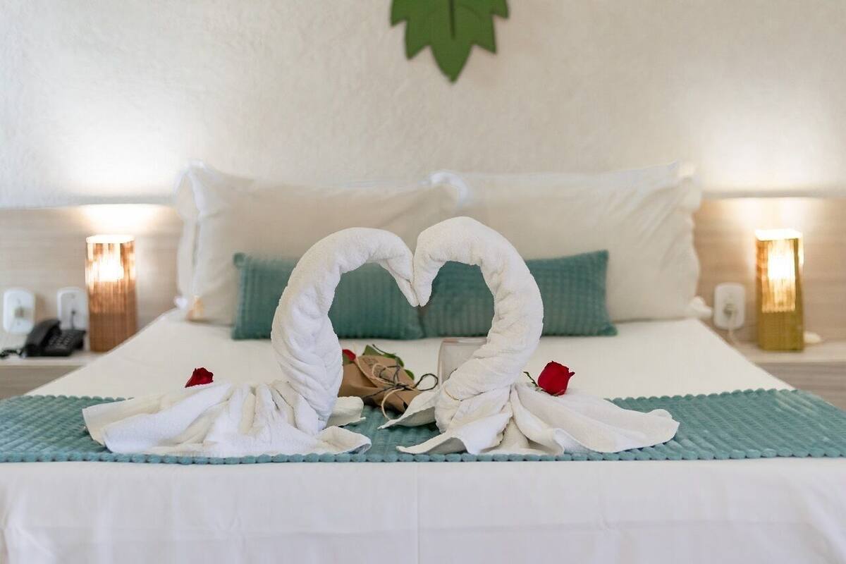 A neatly made bed with a heart-shaped towel arrangement, a bedside table with a lamp, and a wall-mounted telephone.