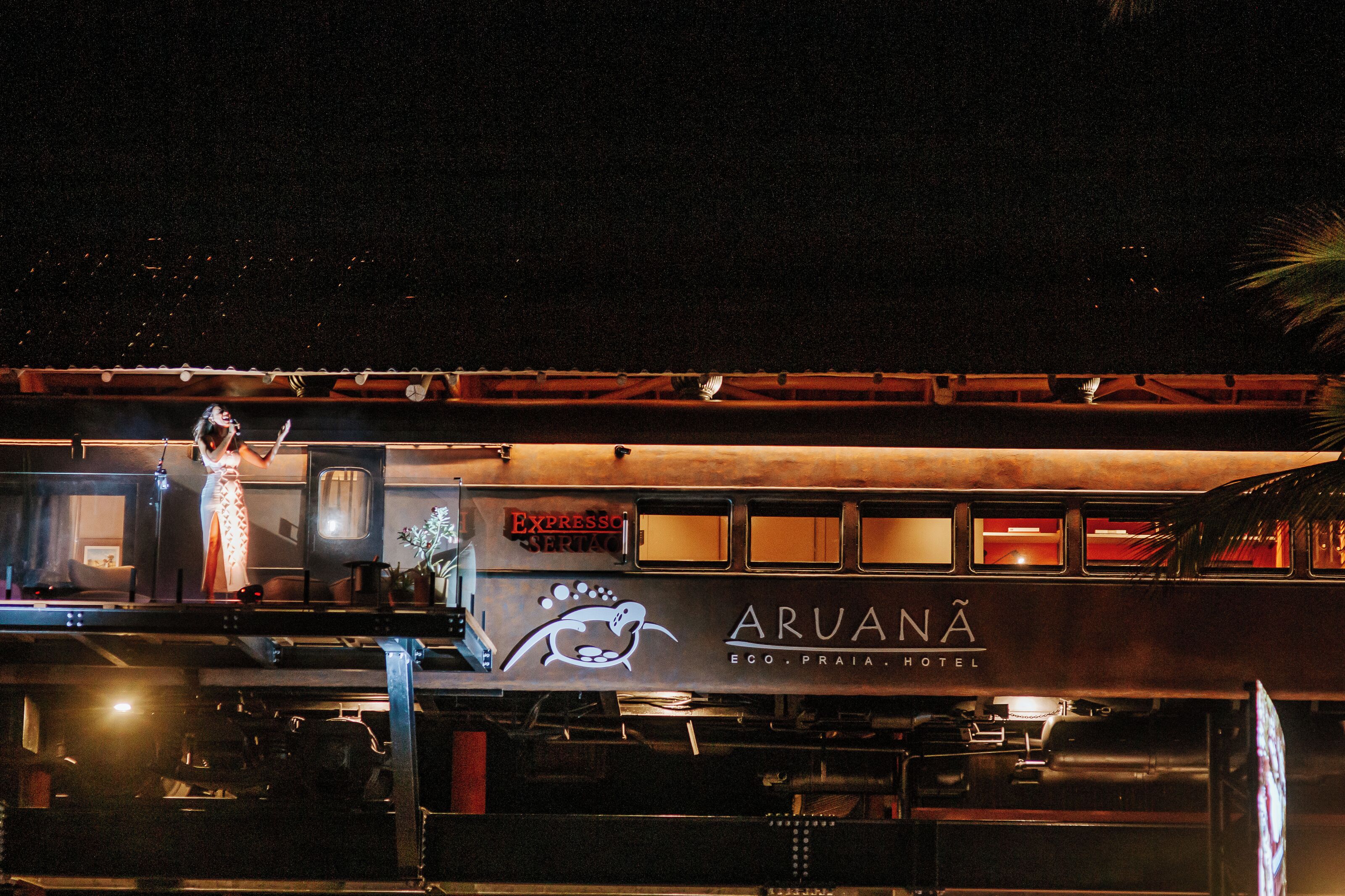 Ein stehender Zug mit der Beschriftung Aruana Eco, Praia Hotel, einem Speisewagen und einem Bahnsteig, auf dem sich eine Person befindet.