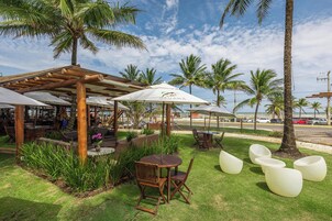An outdoor seating area with wooden tables and chairs, surrounded by palm trees and a view of the beach.