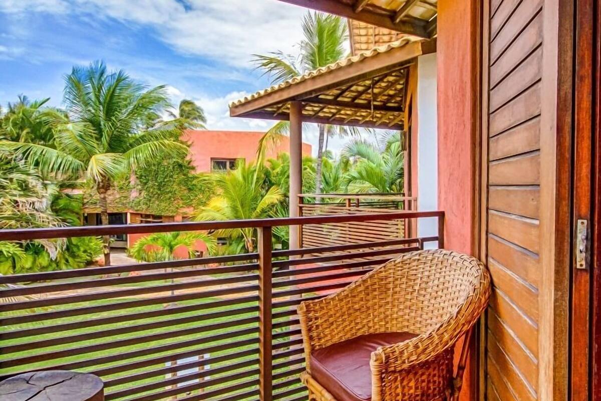 A balcony with a wicker chair, stone flooring, and a view of palm trees and a building in the background.
