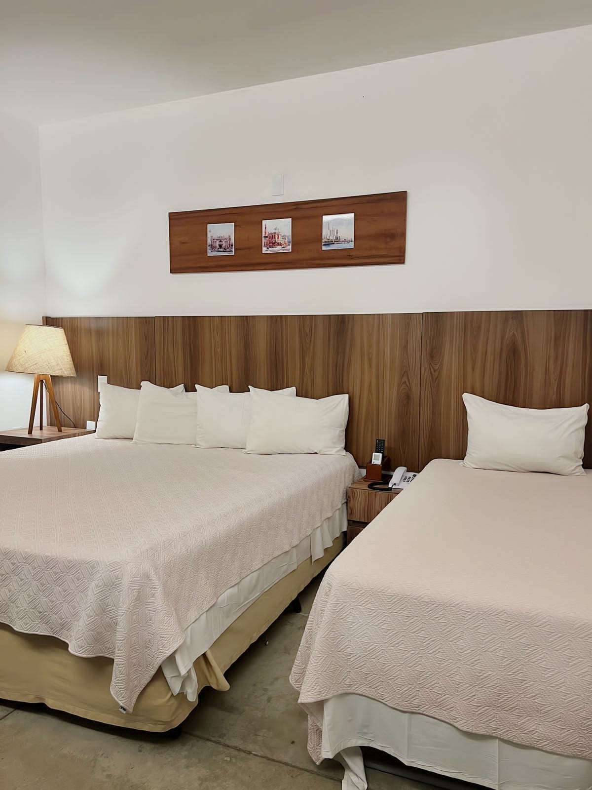 Habitación de hotel con dos camas, cabecero de madera, mesita de noche con lámpara y un cuadro colgado en la pared.